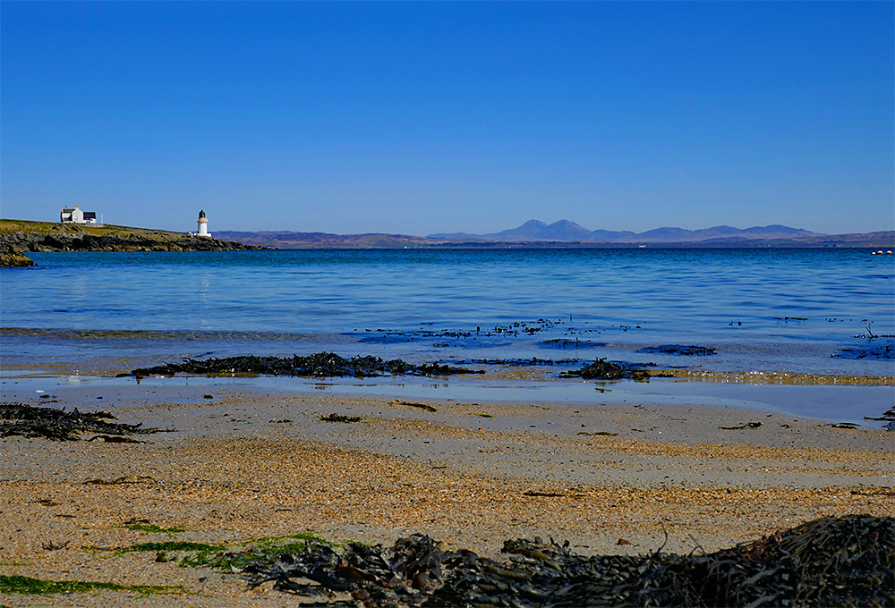 Picture of a view from a sandy beach over a sea loch, a small lighthouse on the shore to the left, some mountains on the horizon