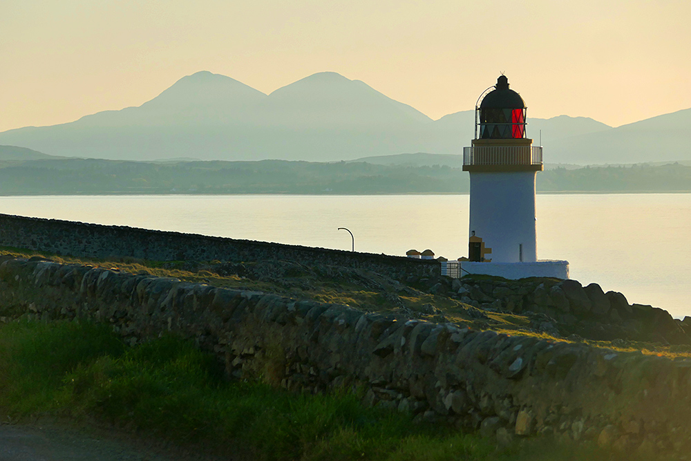 Picture of a small lighthouse in the April morning light, two round mountains in the hazy background