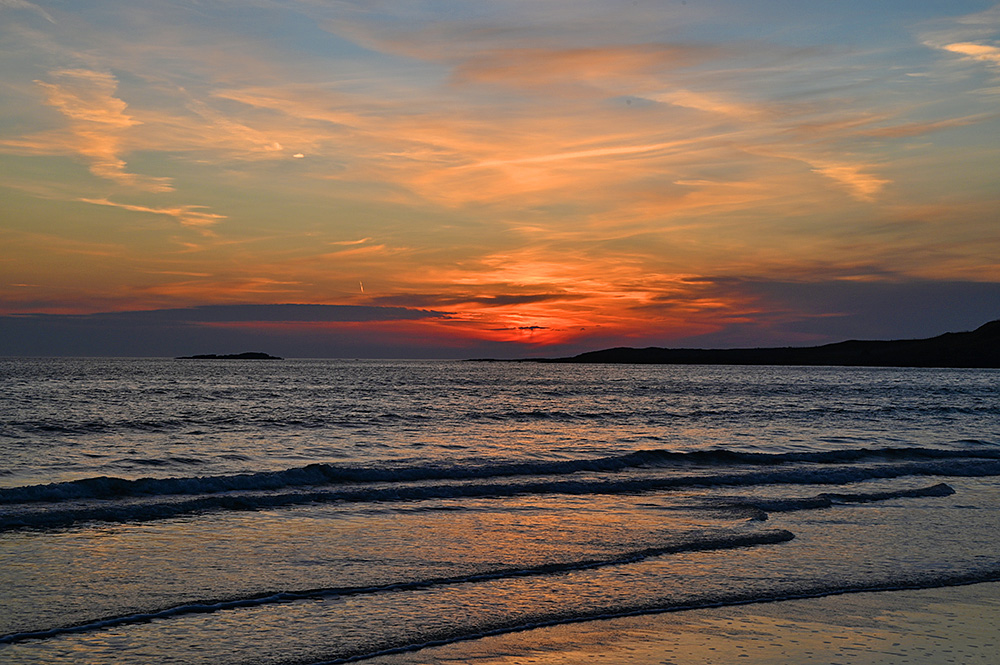 Picture of a cloudy but still colourful sunset seen from a sandy beach with small waves rolling up