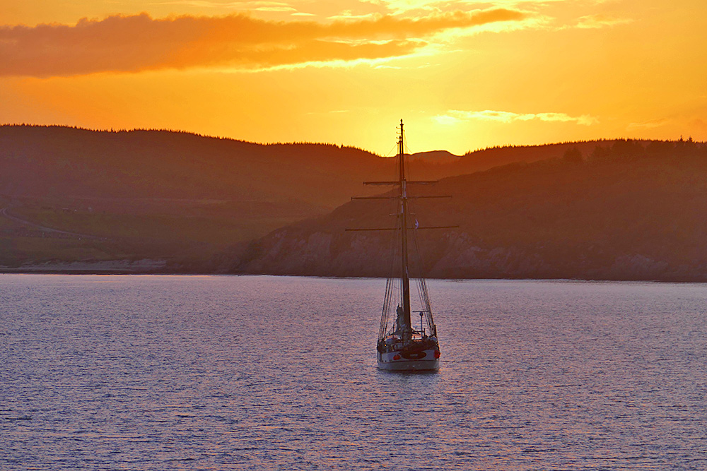 Picture of a small tall ship anchored in a wide bay at sunset, the sun just setting behind some low hills in the background