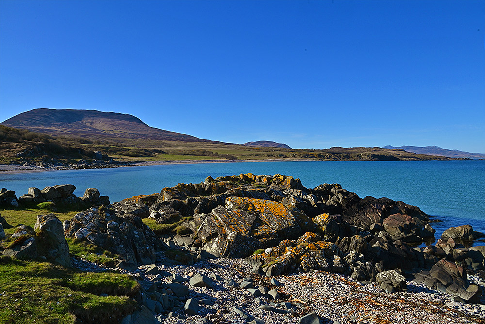 Picture of a wide bay with a pebble beach, a mountain in the background, seen across a rocky shore
