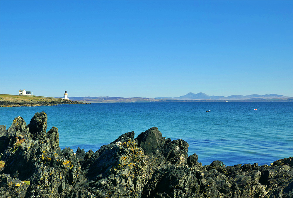 Picture of jagged rocks at the shore of a sea loch, a small lighthouse and some round mountains in the distance