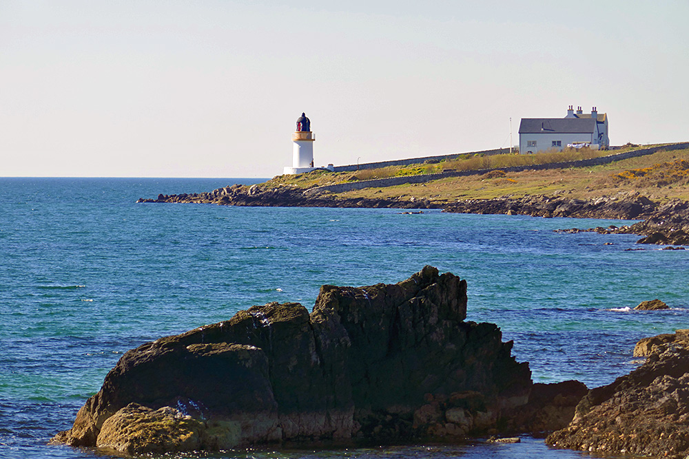 Picture of a small lighthouse on a rocky shoreline, a house with the washing out on the line also visible