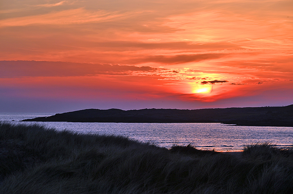 Picture of the setting sun disappearing behind clouds over cliffs, seen from a path through dunes