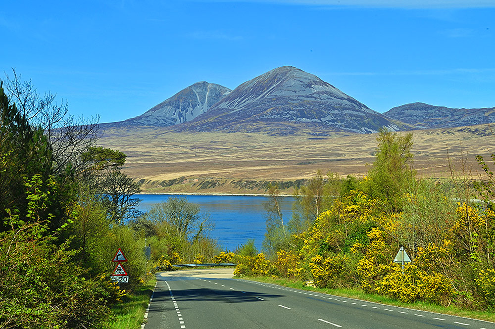 Picture of a road leading down a hillside to a sound between two islands, two mountains covered in scree on the other island