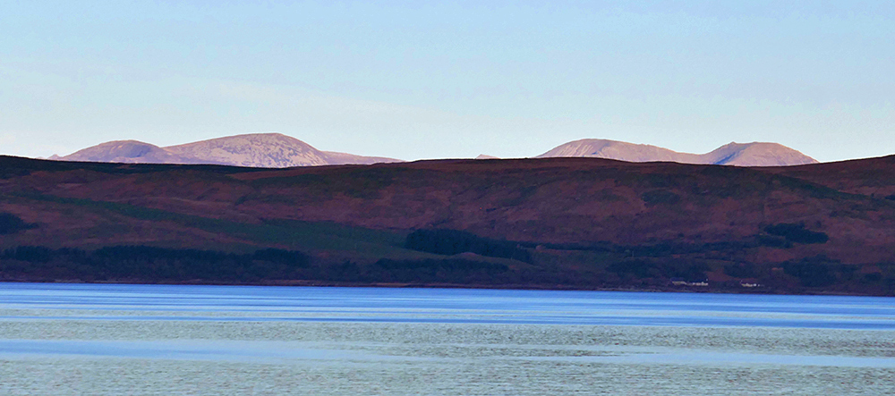 Picture of the bright tops of some distant hills, seen behind some darker lower hills. Viewed across water
