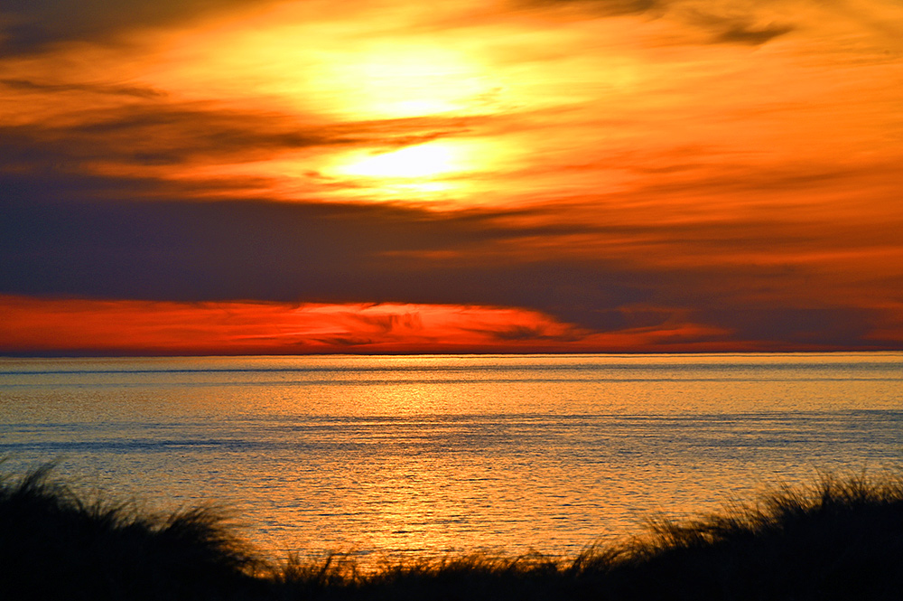 Picture of a colourful cloudy sunset over a wide bay, seen from dunes with the dune grass just visible