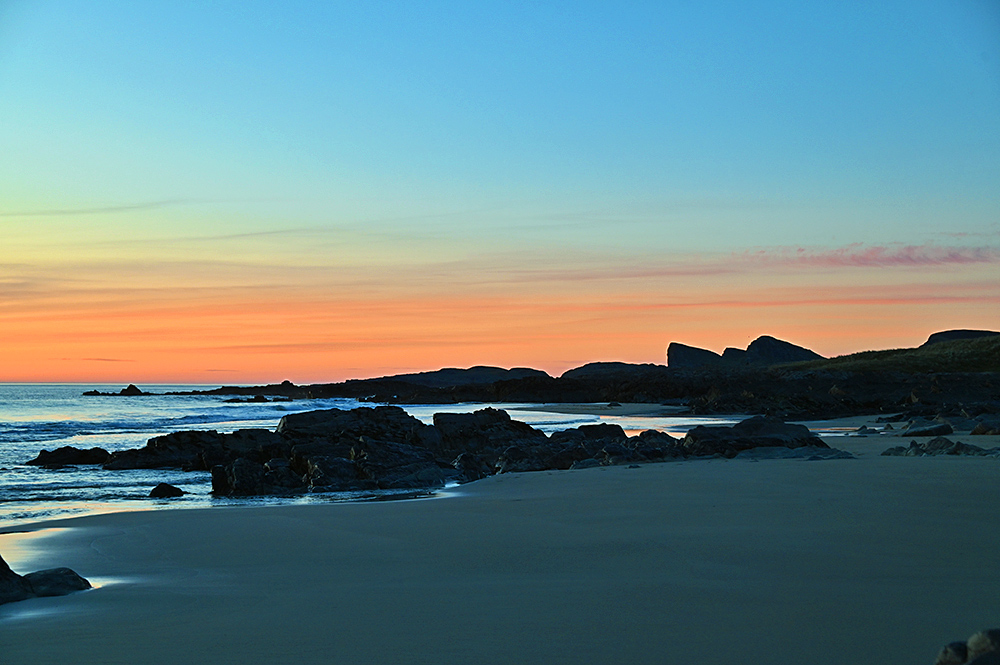 Picture of a colourful sky during the gloaming over a bay with a beach and rocky outcrops