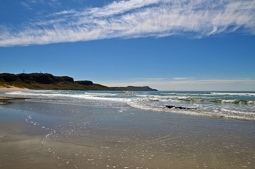 Picture of a long sandy beach in a wide bay with the remains of a wreck along the waterline and some interesting fluffy clouds above