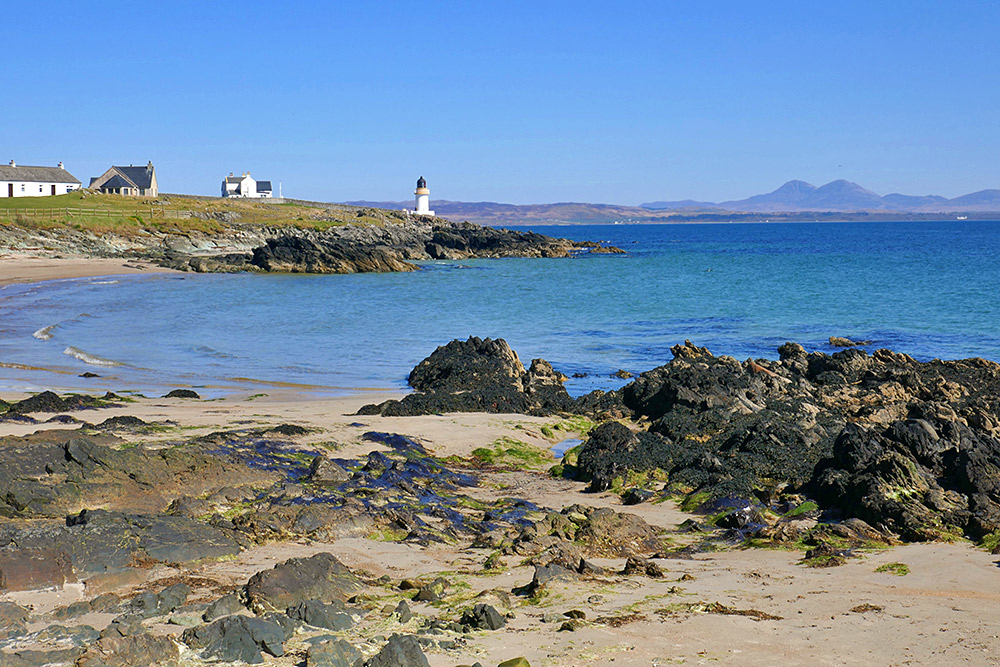 Picture of a view from a small beach towards a small lighthouse and across a sea loch to distant mountains