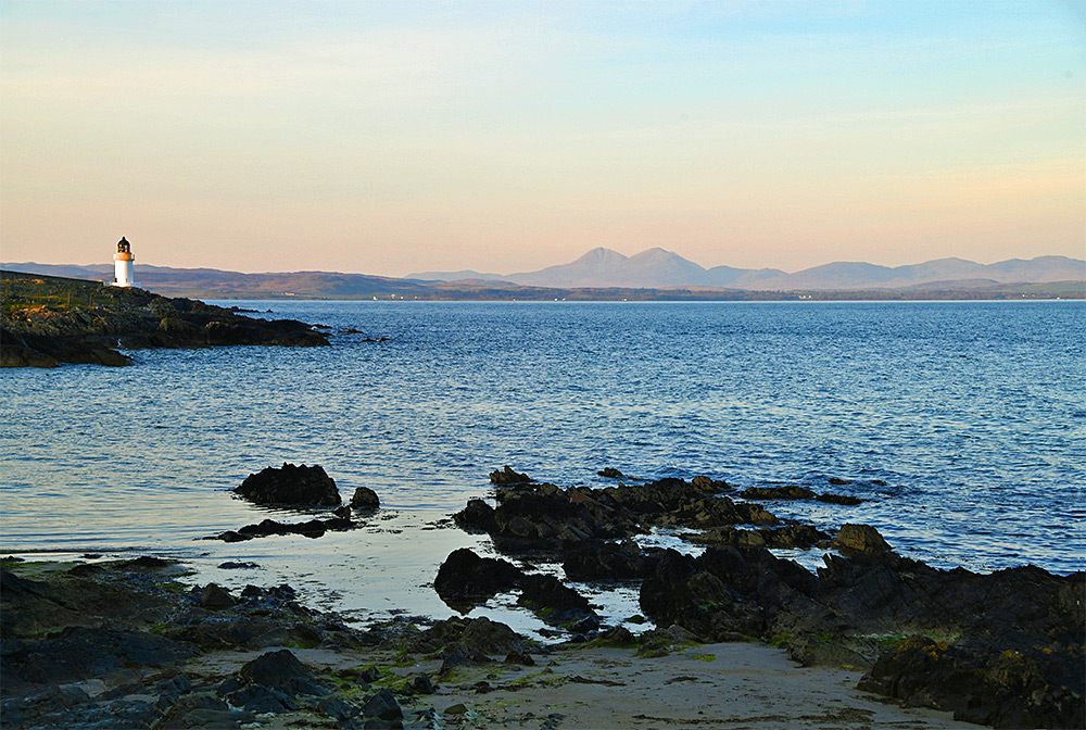 Picture of a view from a small beach over a sea loch with a small lighthouse and some mountains in the distance