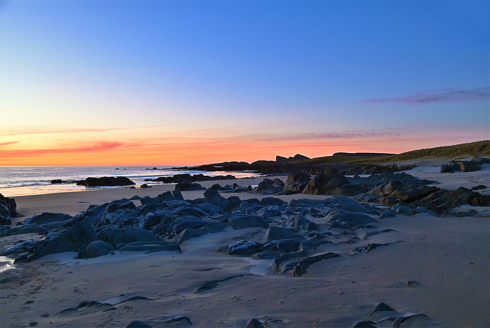 Picture of a beach with rugged rocks embedded in the sand under the mild light of the gloaming