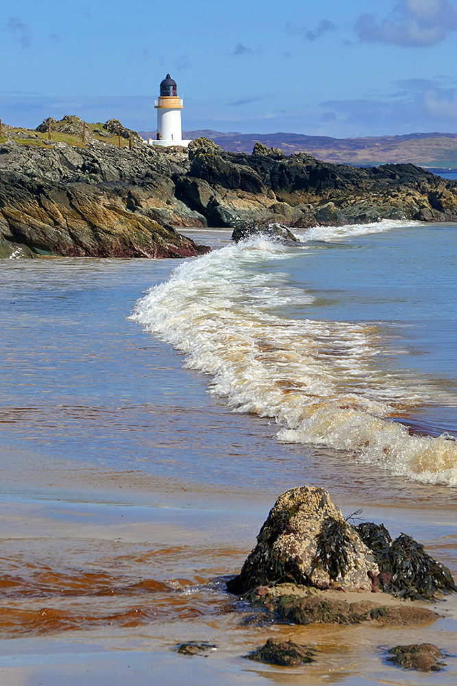 Picture of a wave rolling out on a small beach, a small lighthouse on a rocky shore behind it