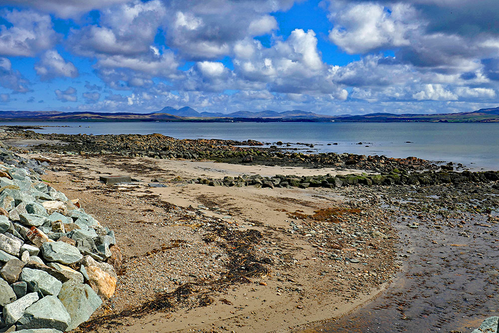 Picture of a rocky shoreline at a sea loch, mountains in the distance on the other side of the loch. Some clouds overhead