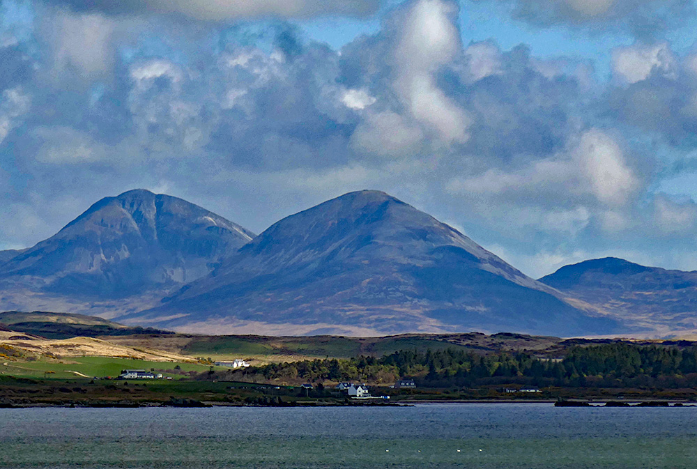 Picture of two mountains towering over a small coastal settlement, some rocks in the water off the shore