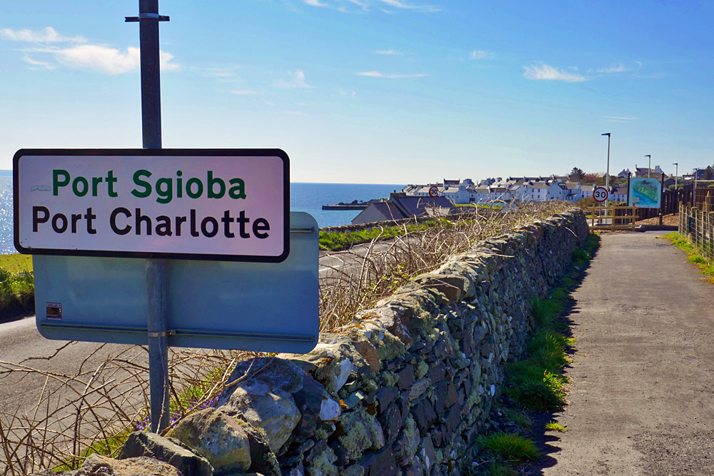 Picture of a path along a stone wall entering a coastal village, a sign reading Port Sgioba / Port Charlotte next to the wall