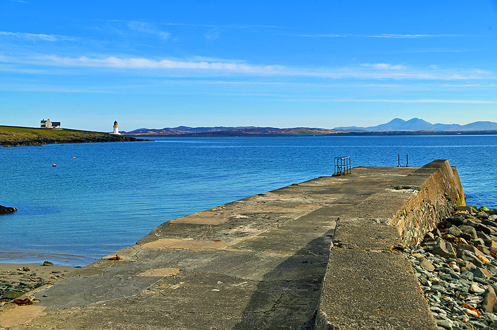 Picture of a small pier at a sea loch, a small lighthouse in the distance, also some mountains in the far distance