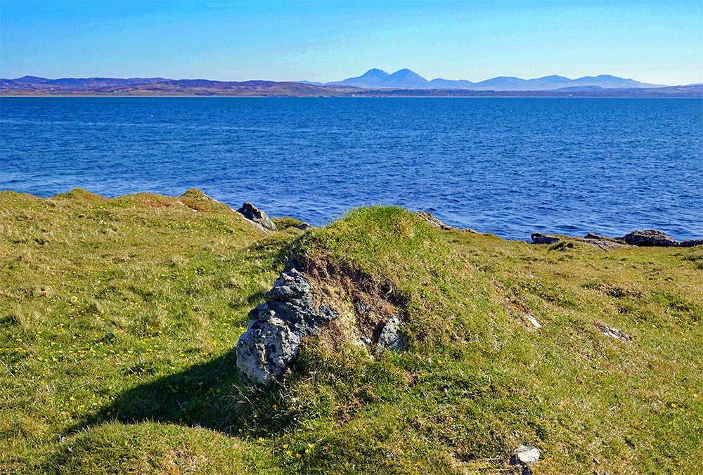 Picture of a large rock with some turf over it on the shore of a sea loch, mountains in the distance across the loch