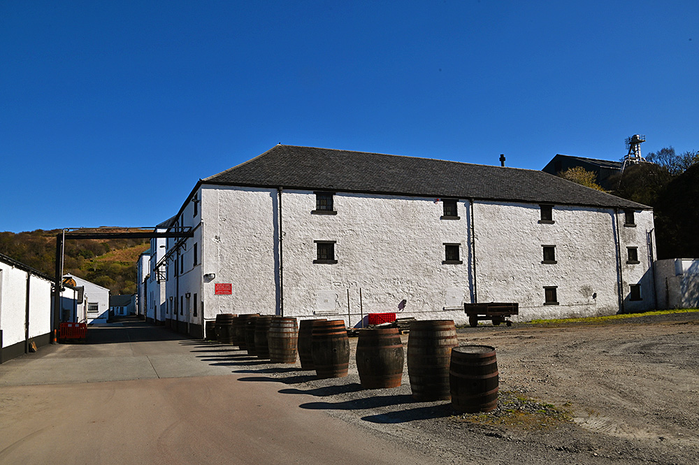 Picture of some whitewashed distillery warehouses under a bright blue sky, some casks in front of them