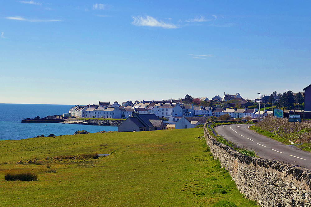 Picture of a coastal village with mostly whitewashed buildings in bright April sunshine, seen from next to the road leading to the village