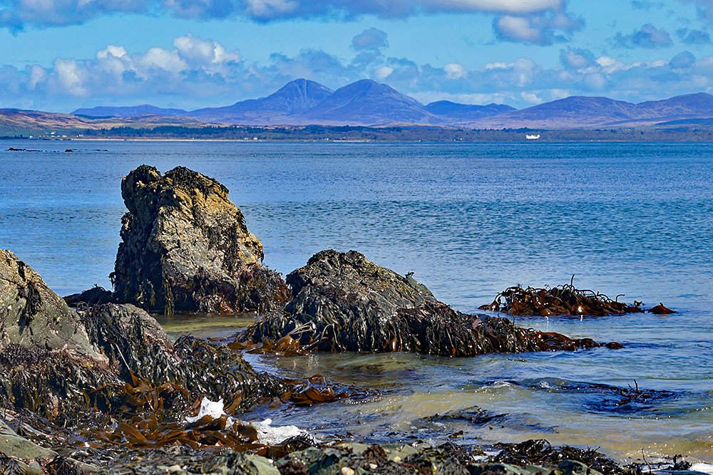 Picture of some rocks at the end of a sea loch beach, mountains in the distance on the other side of the loch