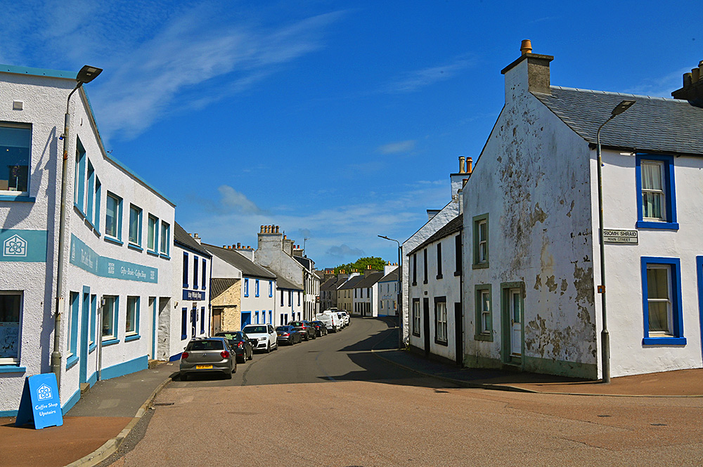Picture of a narrow village street with shops and houses, cars parked on the left