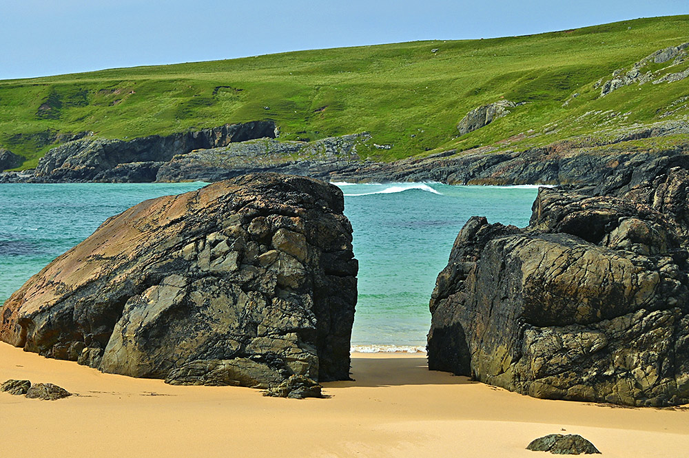 Picture of two big boulders on a sandy beach with a wave breaking behind