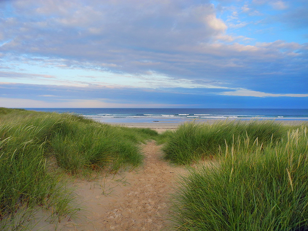 Picture of a view from a path through dunes to a beach under a partially cloudy sky with the morning sun breaking through