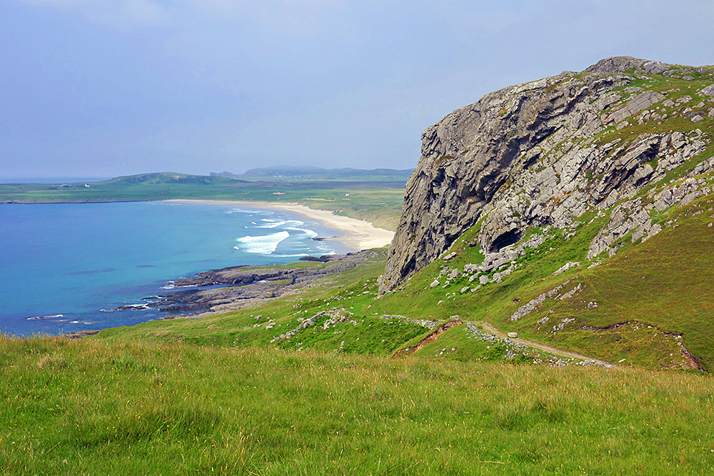 Picture of a view from a small hill past some steep crags to a sandy beach in a wide bay