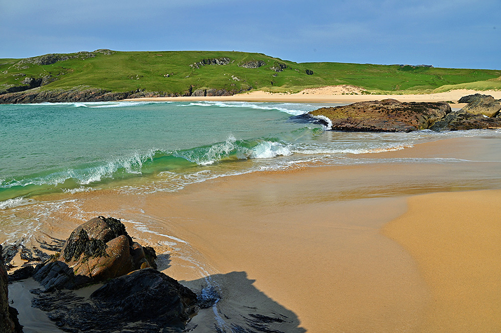 Picture of a small wave cresting and breaking as it reaches a sandy beach in a small bay