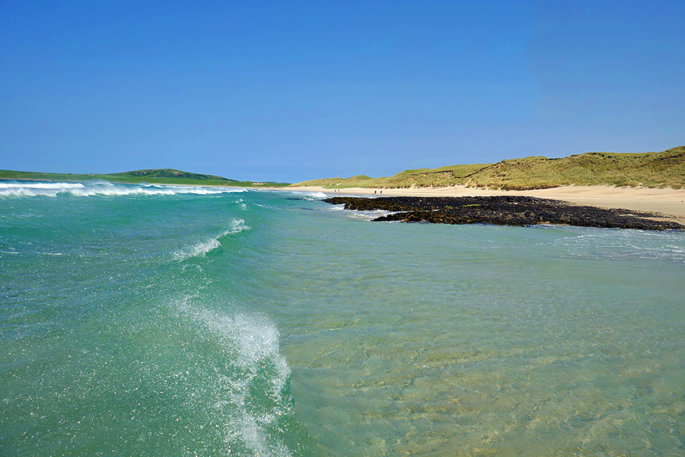 Picture of a small wave breaking and sending spray into the air as it approaches a rocky outcrop on a sandy beach